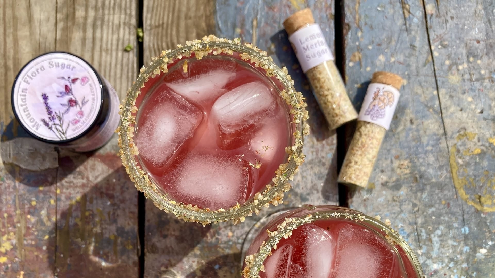 A wooden background with a glass of pink liquid, a vial of Lemon Meringue Pie infused sugar, and a jar of Mountain Flora Sugar from Well Seasoned Table.