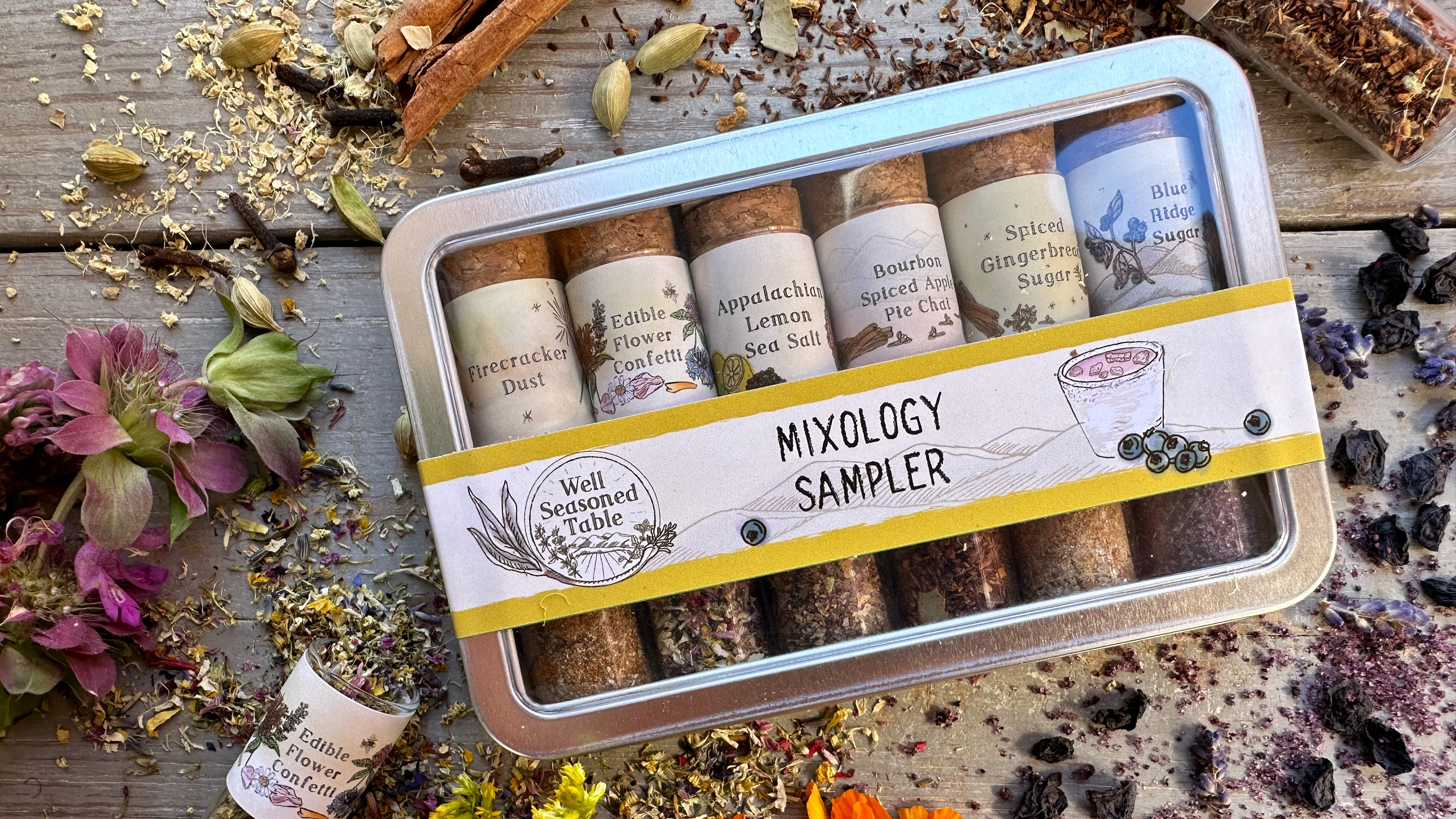 A sampler tin of vials of spices from Well Seasoned Table on a wooden background with organic dried fruit and flowers around it.