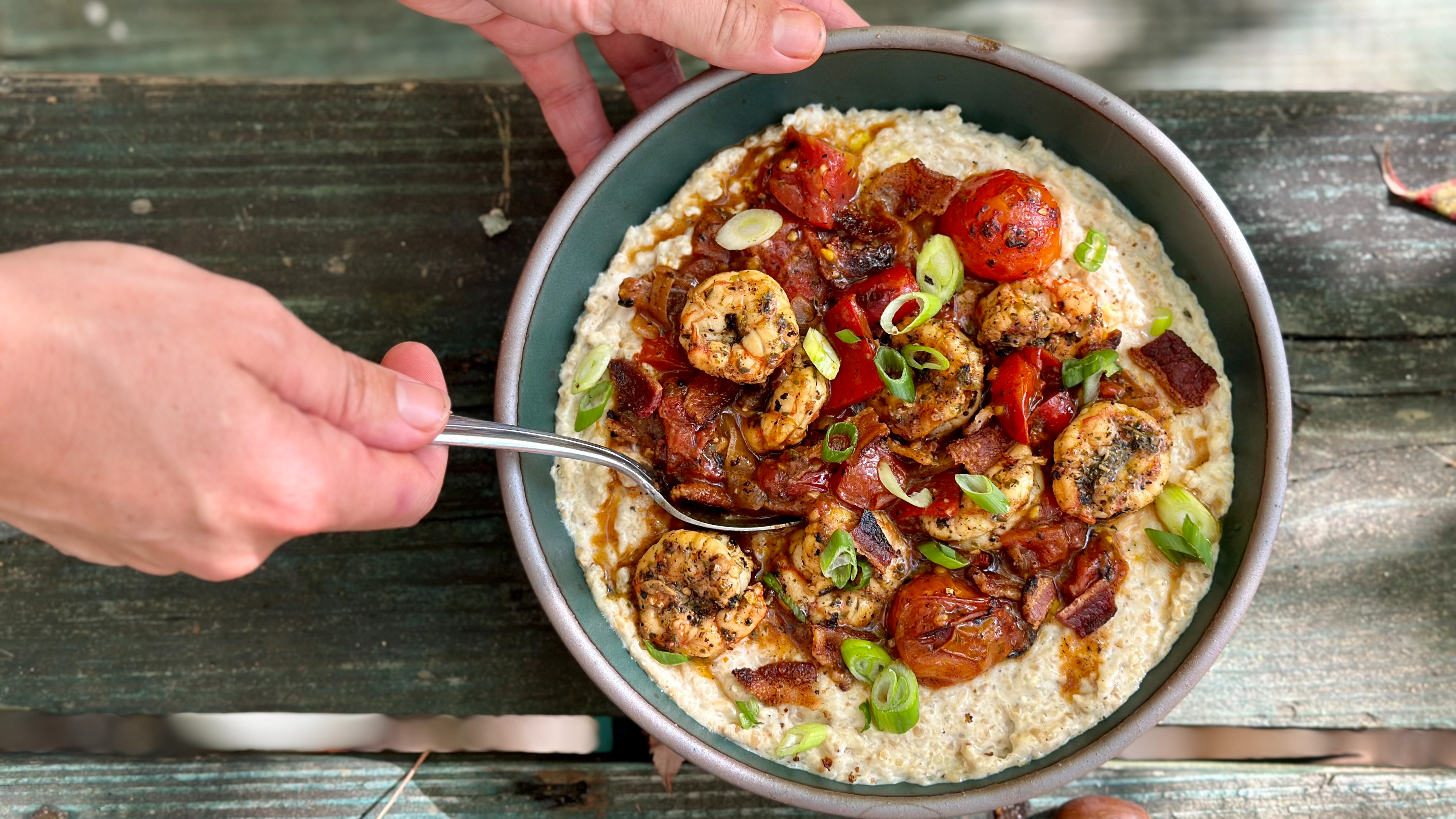 A bowl of creamy-looking grits topped with shrimp and sauce, with hands holding it and a wooden backdrop.