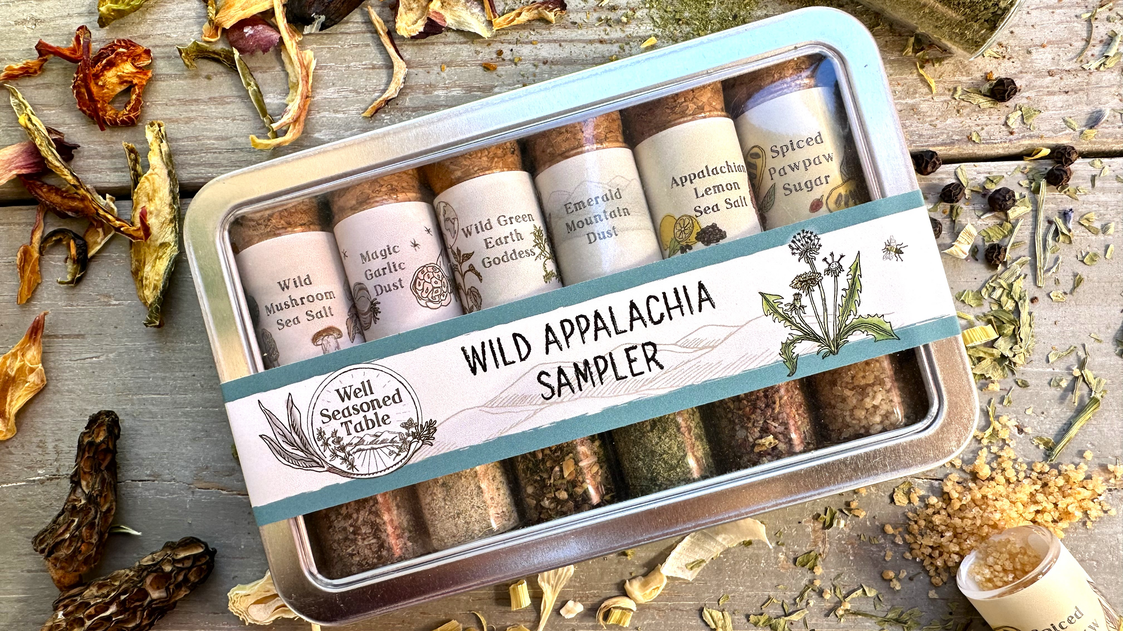 A sampler tin of vials of spices from Well Seasoned Table on a wooden background with organic dried spices around it.