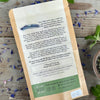 The back of a packet of in the garden with tulsi tea from Well Seasoned Table on a wooden background with fresh spearmint and cornflowers around it and a ceramic bowl of tea.