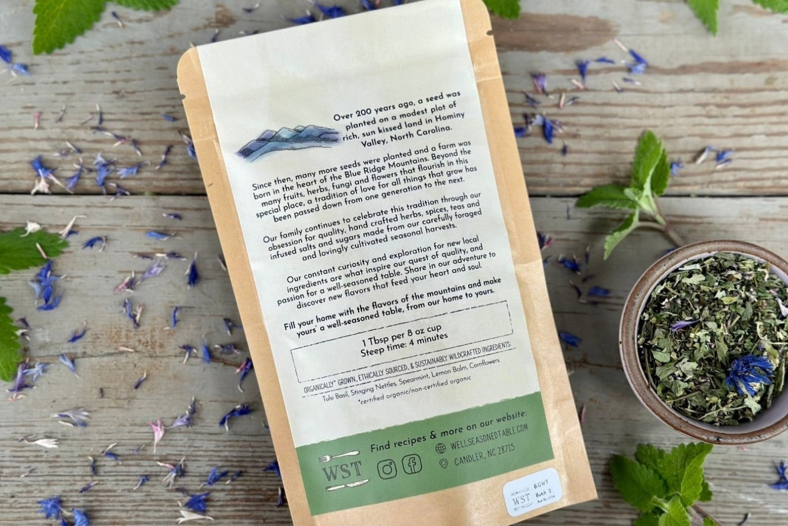 The back of a packet of in the garden with tulsi tea from Well Seasoned Table on a wooden background with fresh spearmint and cornflowers around it and a ceramic bowl of tea.