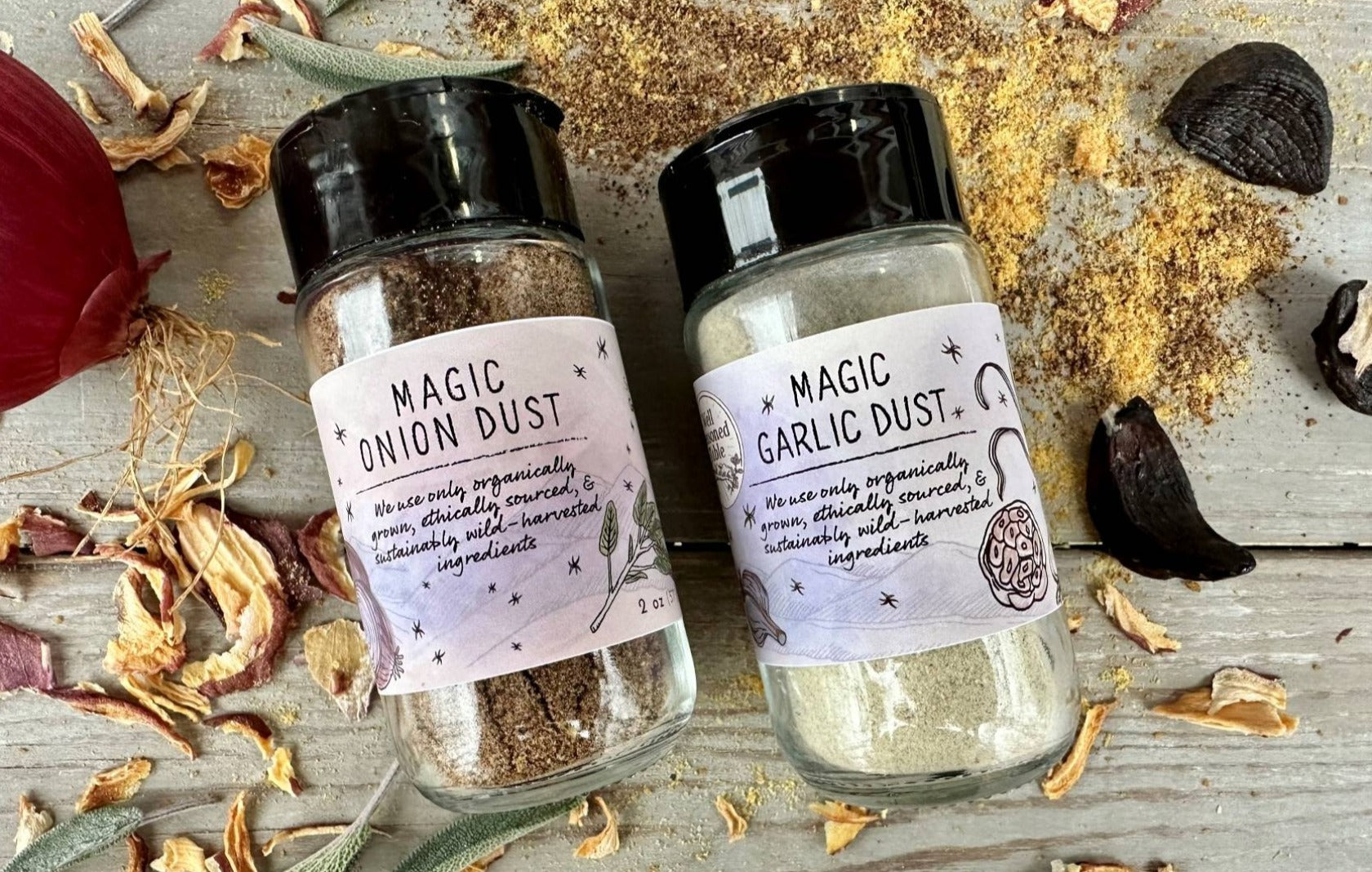 A glass seasoning jar of Magic Garlic Dust and Magic Onion Dust from Well Seasoned Table on a wooden background with onions, sage, and black garlic around it.