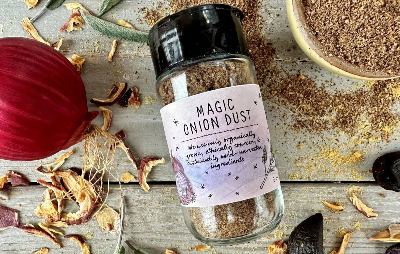 Close up of a glass seasoning jar of Magic Onion Dust from Well Seasoned Table on a wooden background with onions, sage, and black garlic around it.