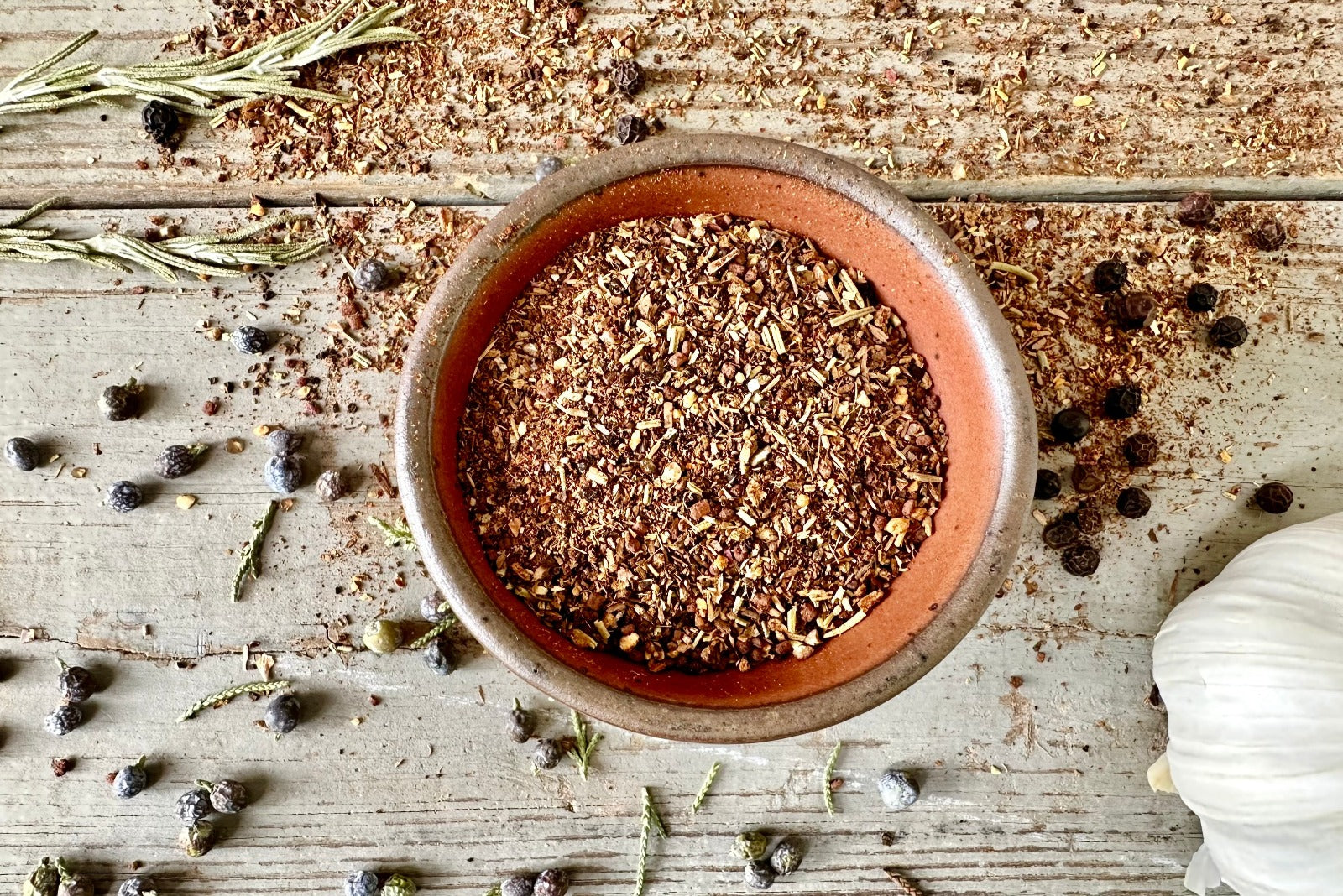 A bowl of Smoky Mountains Grilling Rub from Well Seasoned Table on a wooden background with a bowl of seasoning, juniper berries, black peppercorns, garlic, and rosemary.