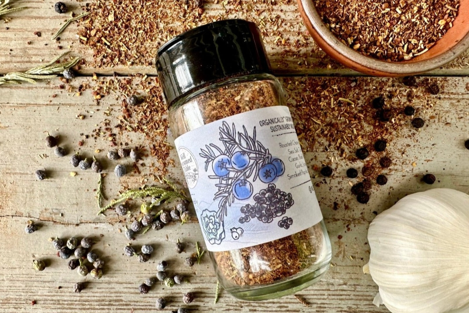 The side of a glass shaker jar of Smoky Mountains Grilling Rub from Well Seasoned Table on a wooden background with a bowl of seasoning, juniper berries, black peppercorns, garlic, and rosemary.