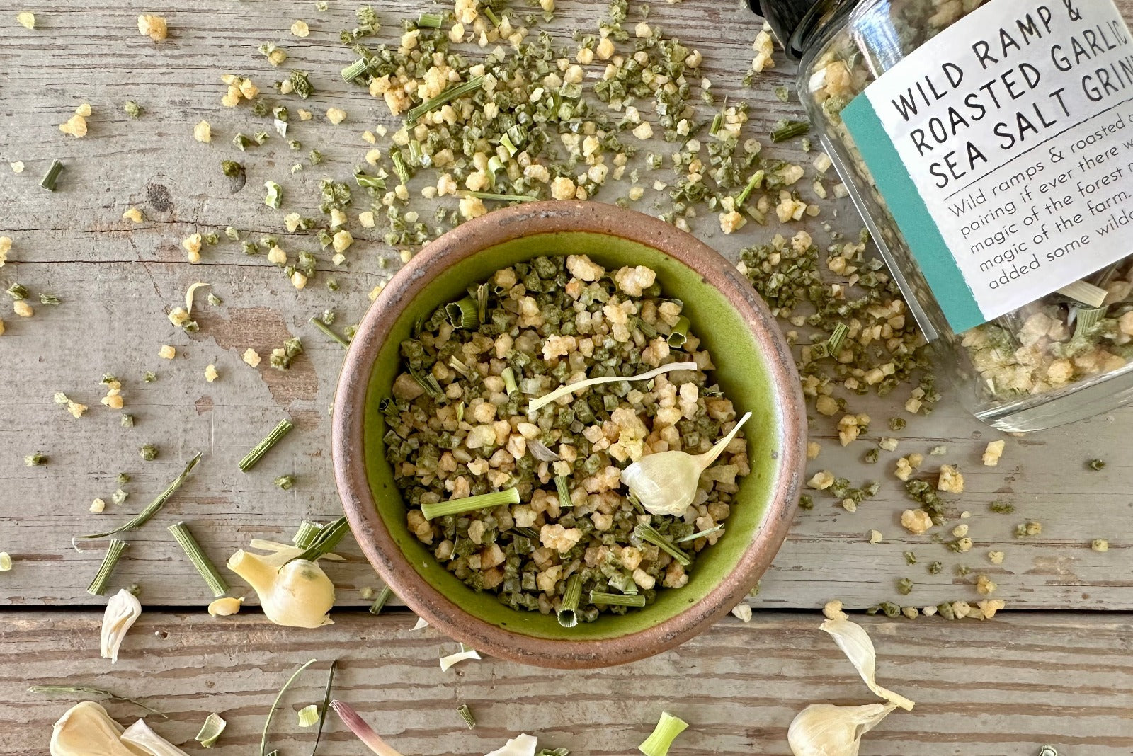 A bowl of Wild Ramp and Roasted Garlic Sea Salt from Well Seasoned Table next to a grinder jar and on a wooden background with dried wild garlic.
