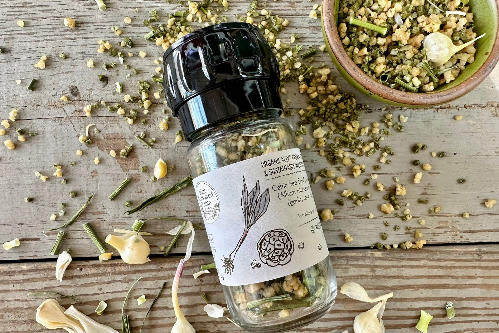 The side of a glass grinder jar of Wild Ramp and Roasted Garlic Sea Salt from Well Seasoned Table on a wooden background with a bowl of product and some dried wild garlic around it.