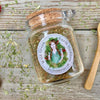A Glass jar with cork and label and a small wooden spoon on a wooden background with dried herbs, garlic, and sumac around it.