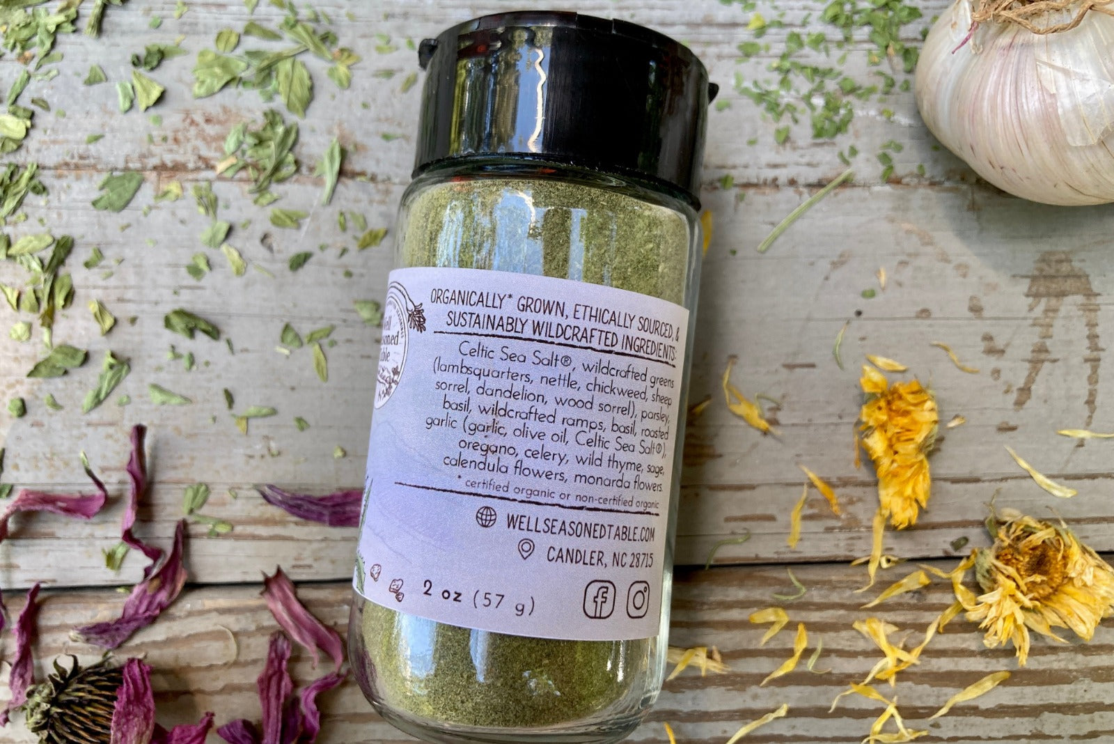 A jar of Emerald Mountain Dust from Well Seasoned Table on a wooden background with a sprinkle of calendula flowers and dried ramps.