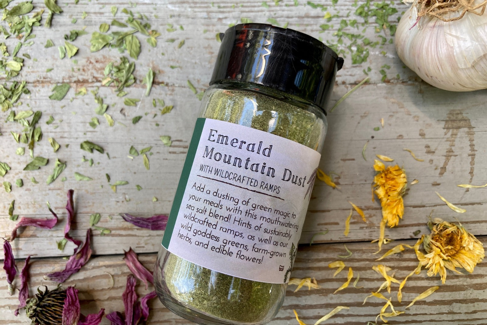 A jar of Emerald Mountain Dust from Well Seasoned Table on a wooden background with a sprinkle of calendula flowers and dried ramps.