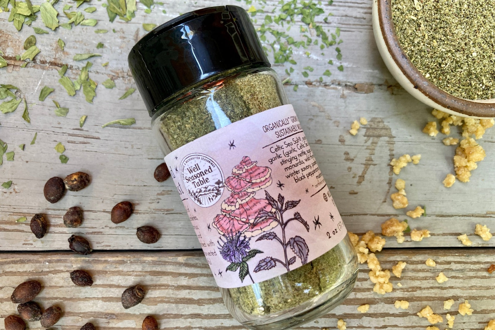 A glass shaker jar of Enchanted Forest dust from Well Seasoned Table, a wild mushroom and herb infused sea salt, on a wooden background with garlic, herbs, ramps, and spicebush berries.