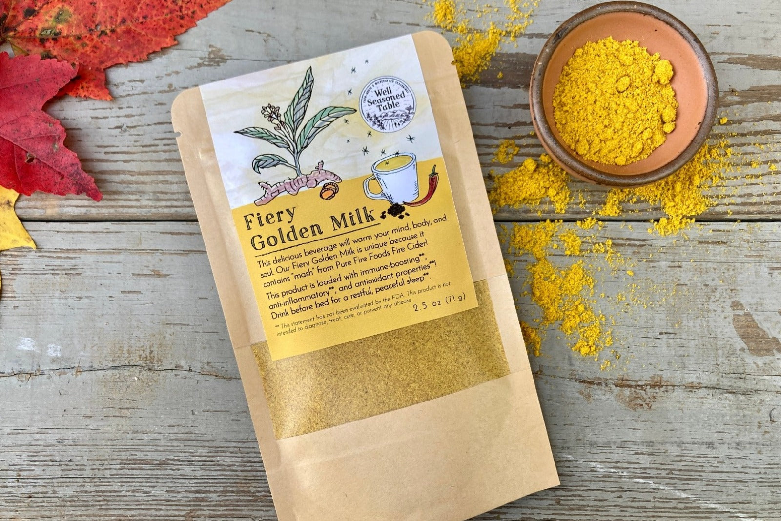 A packet of Fiery Golden Milk from Well Seasoned Table on a wooden background with fall leaves and a bowl of golden milk powder.