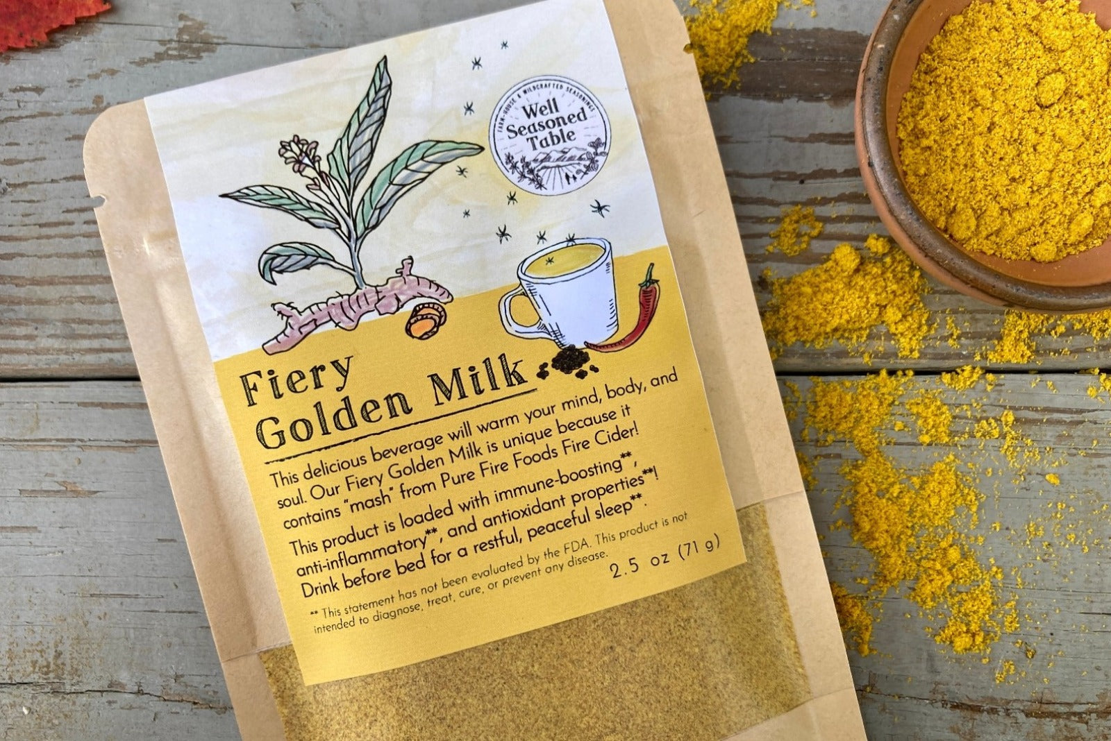 A packet of Fiery Golden Milk from Well Seasoned Table on a wooden background with fall leaves and a bowl of golden milk powder.