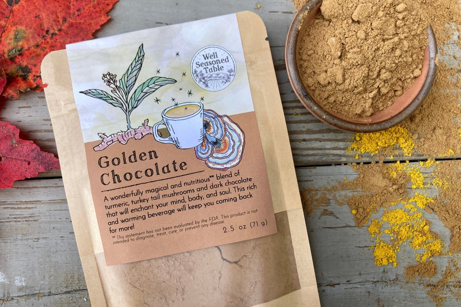 A packet of Golden Chocolate from Well Seasoned Table on a wooden background with a bowl of cocoa powder and a sprinkle of turmeric.