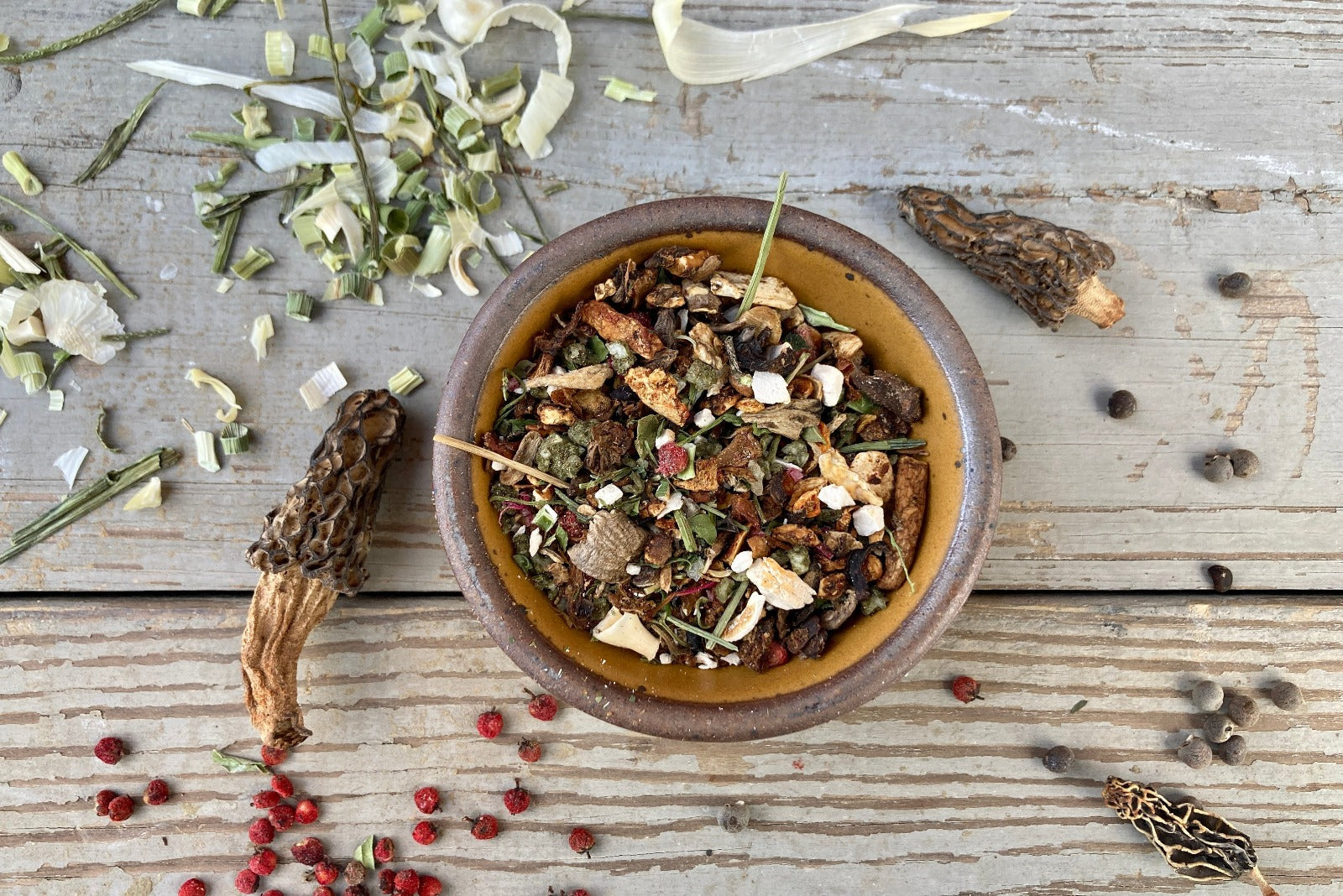 A bowl of Magical Forest Grinder from Well Seasoned Table on a wooden background with wild onions, dried morels, and sumac berries around it.