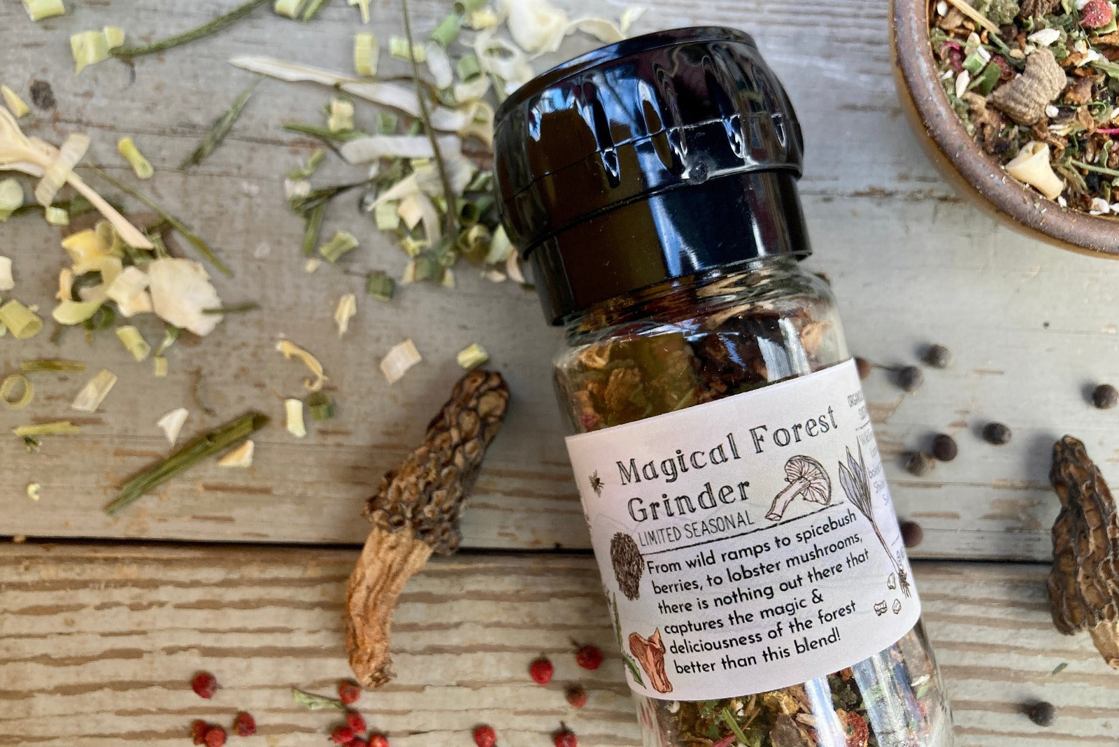 A glass grinder jar of Magical Forest Grinder from Well Seasoned Table on a wooden background with wild onions, dried morels, and sumac berries around it.