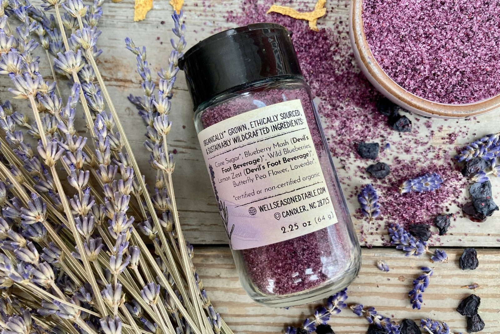 The back of a glass shaker jar of Blue Ridge Sugar from Well Seasoned Table on a wooden background with dried lavendar, lemon, and blueberries around it.