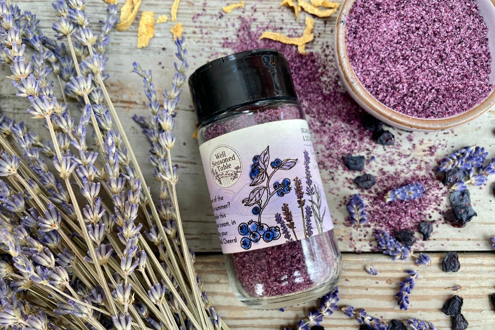 The side of a glass shaker jar of Blue Ridge Sugar from Well Seasoned Table on a wooden background with dried lavendar, lemon, and blueberries around it.