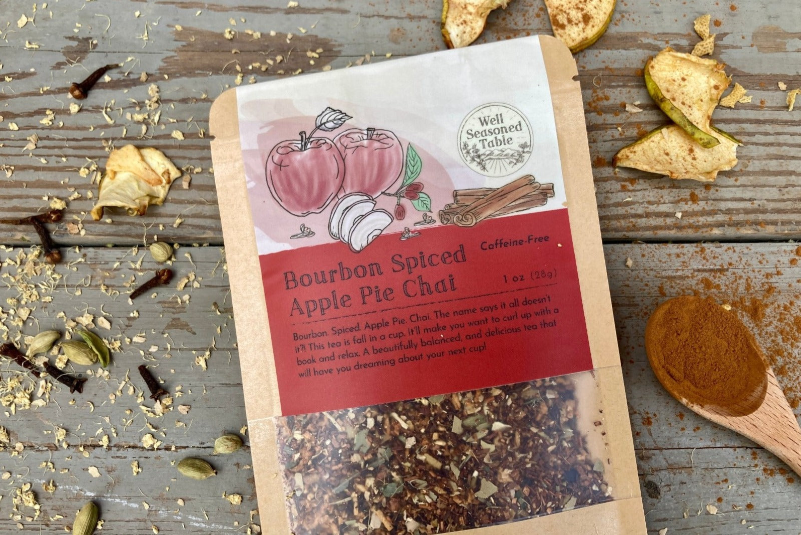 Up close of a packet of Bourbon Spiced Apple Pie Chai Tea from Well Seasoned Table on a wooden background with dried apples, spices, and a spoon of cinnamon around it.