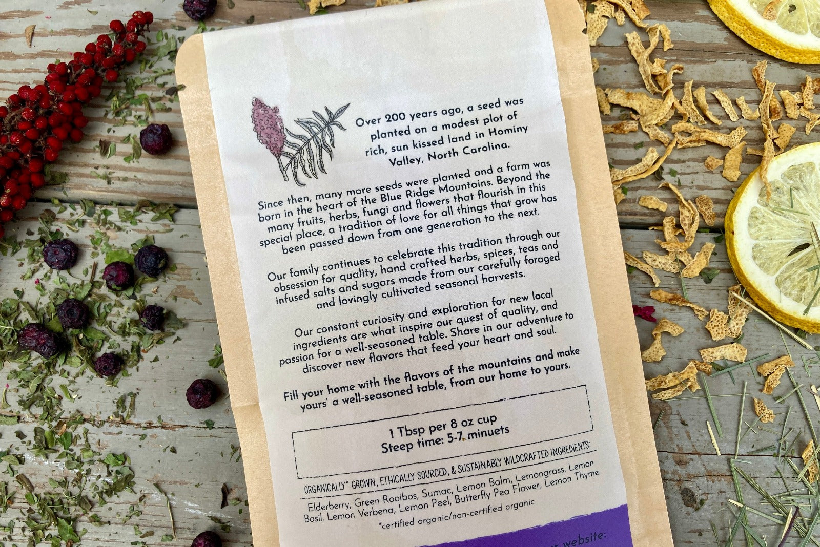 The back of a packet of Elderberry Lemon Tea from Well Seasoned Table on a wooden background with sumac, elderberries, dried lemongrass and lemon slices around it.