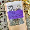 A packet of Elderberry Lemon Tea from Well Seasoned Table on a wooden background with sumac, elderberries, dried lemongrass and lemon slices around it.