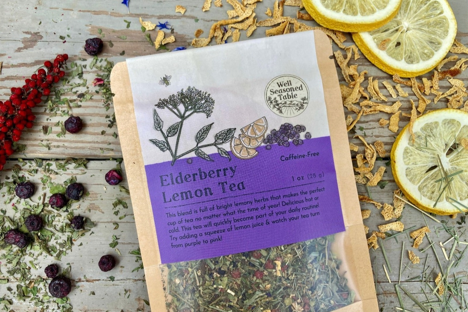 Up close of a packet of Elderberry Lemon Tea from Well Seasoned Table on a wooden background with sumac, elderberries, dried lemongrass and lemon slices around it.
