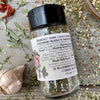 The back of a glass shaker jar of Spice of Life (everything) Seasoning from Well Seasoned Table on a wooden background with a bowl of seasoning, dried peppers, garlic, and herbs.
