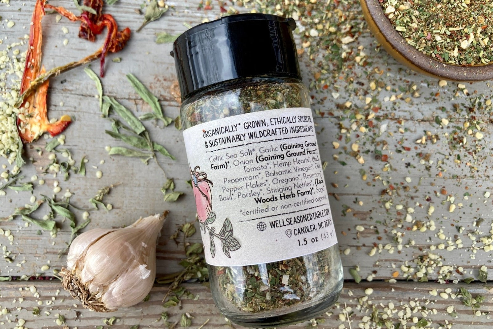 The back of a glass shaker jar of Spice of Life (everything) Seasoning from Well Seasoned Table on a wooden background with a bowl of seasoning, dried peppers, garlic, and herbs.
