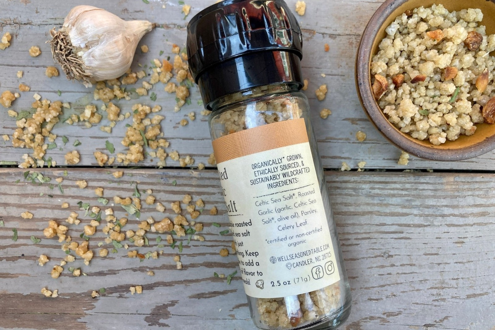 The side of a glass grinder jar of Roasted Garlic Sea Salt on a wooden background with a bowl of the infused sea salt from Well Seasoned Table.