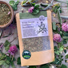 A packet of Wild Green Goddess Seasoning from Well Seasoned Table on a wooden background with wild greens and chive blossoms around it.