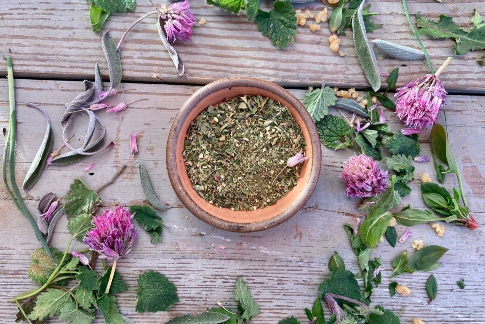 A bowl of Wild Green Goddess Seasoning from Well Seasoned Table on a wooden background with wild greens and chive blossoms around it.
