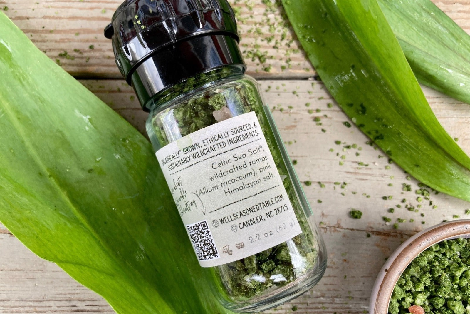 The side of a glass jar of Wild Ramp Sea Salt in a grinder jar from Well Seasoned Table on a wooden background with a small bowl of Wild Ramp Sea Salt.