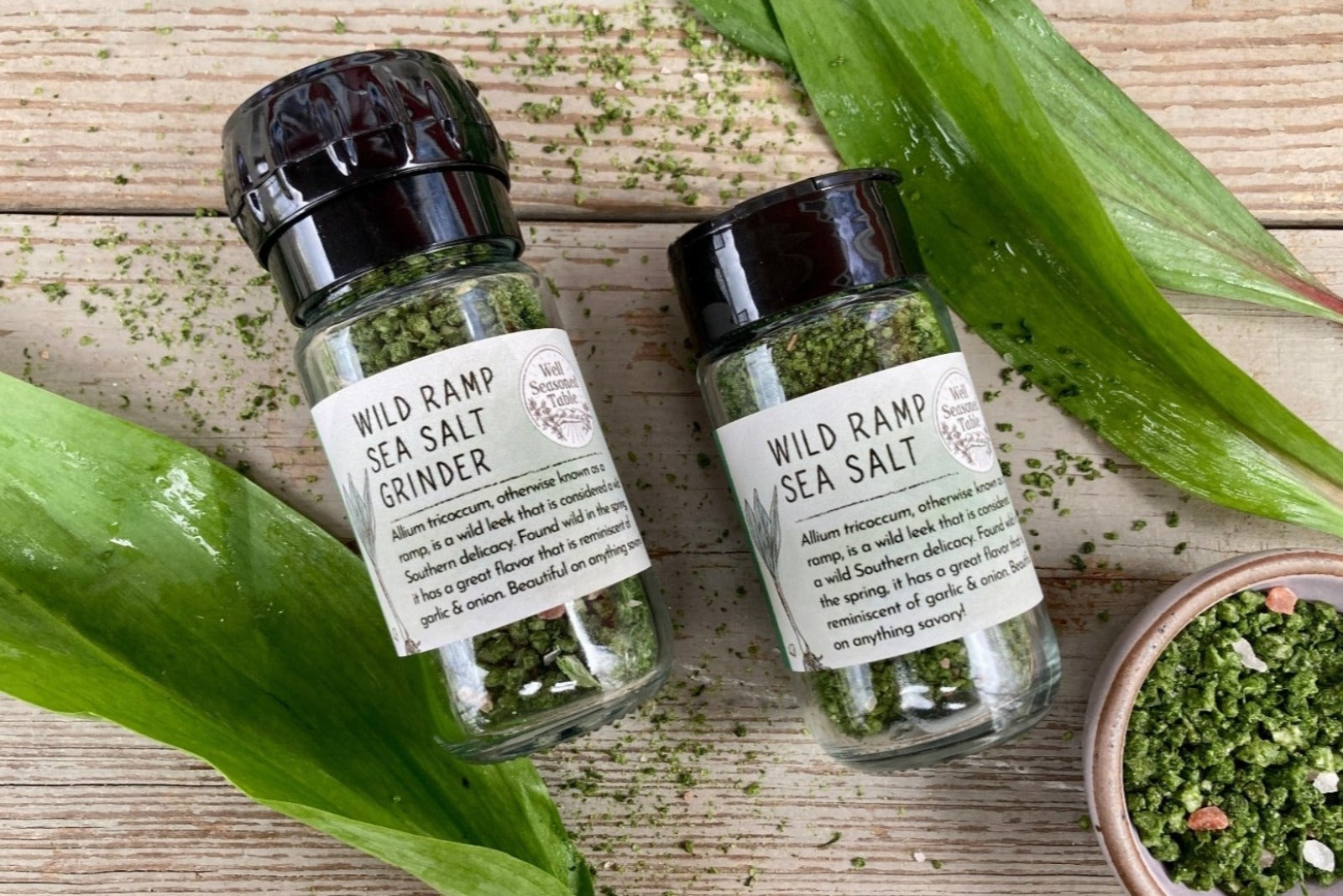 A glass jar of Wild Ramp Sea Salt in a grinder jar and a shaker jar from Well Seasoned Table on a wooden background with a small bowl of Wild Ramp Sea Salt.