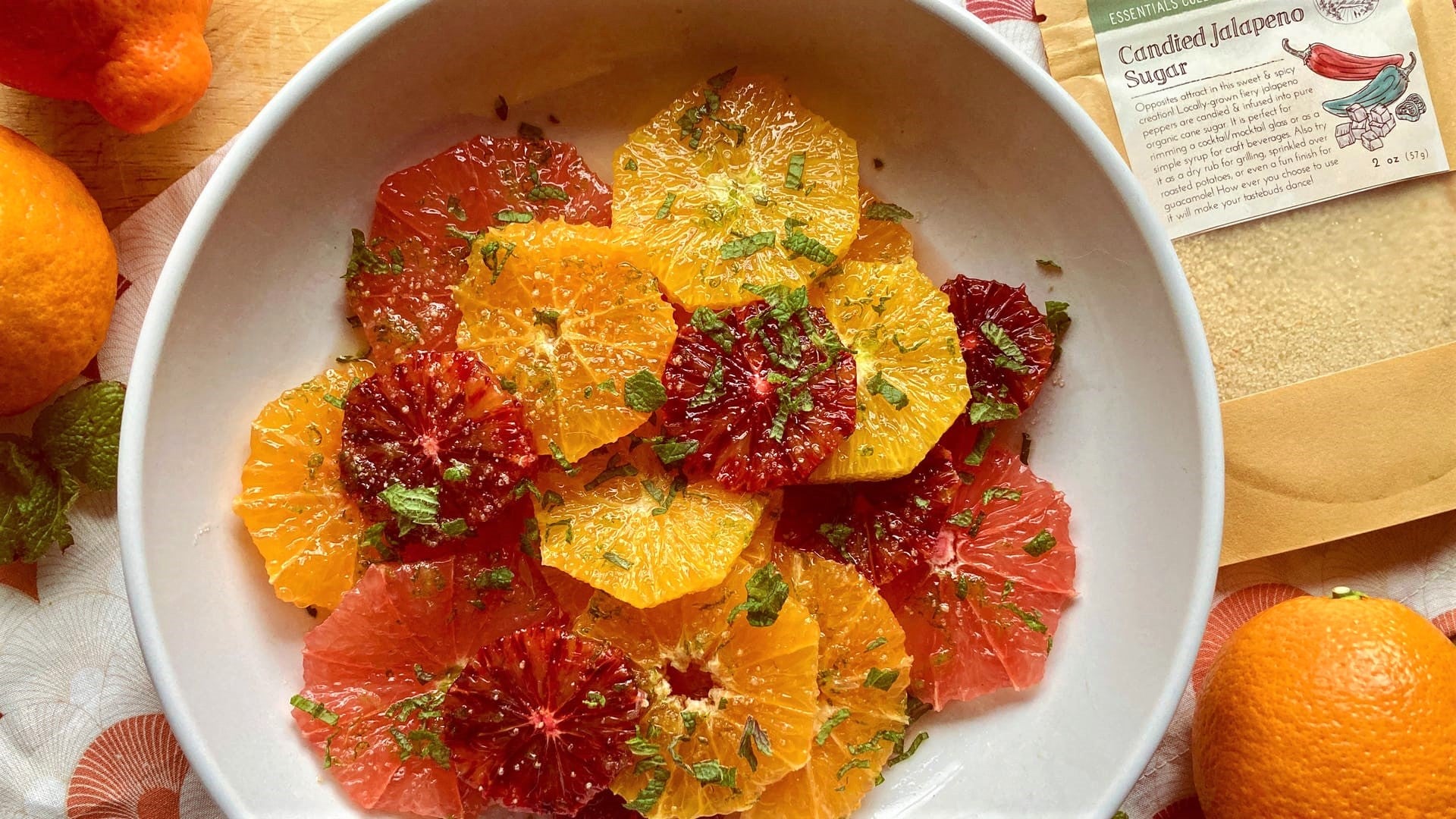 A colorful bowl of citrus slices with a green flecked garnish with citrus fruits in the background and a bag of Well Seasoned Table Candied Jalapeno Sugar