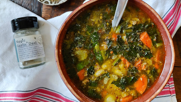 A bowl of colorful soup in a red bowl, on a striped towel, next to an empty jar of Magic Garlic Dust from Well Seasoned Table