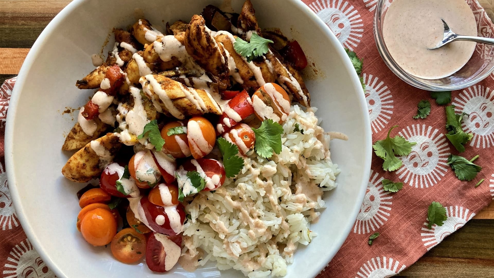 A white bowl of chicken fajitas over rice with cherry tomatoes and Well Seasoned Table organic spices on a tea towel with a bowl of creamy taco seasoned sauce