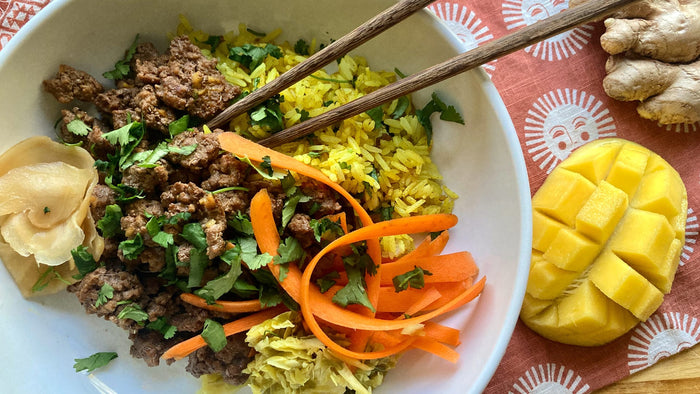 A white bowl with ground beef, ribboned carrots, and yellow rice on a napkin with sun faces, a mango, ginger, and chopsticks. Beef is seasoned with Well Seasoned Table organic spices. 