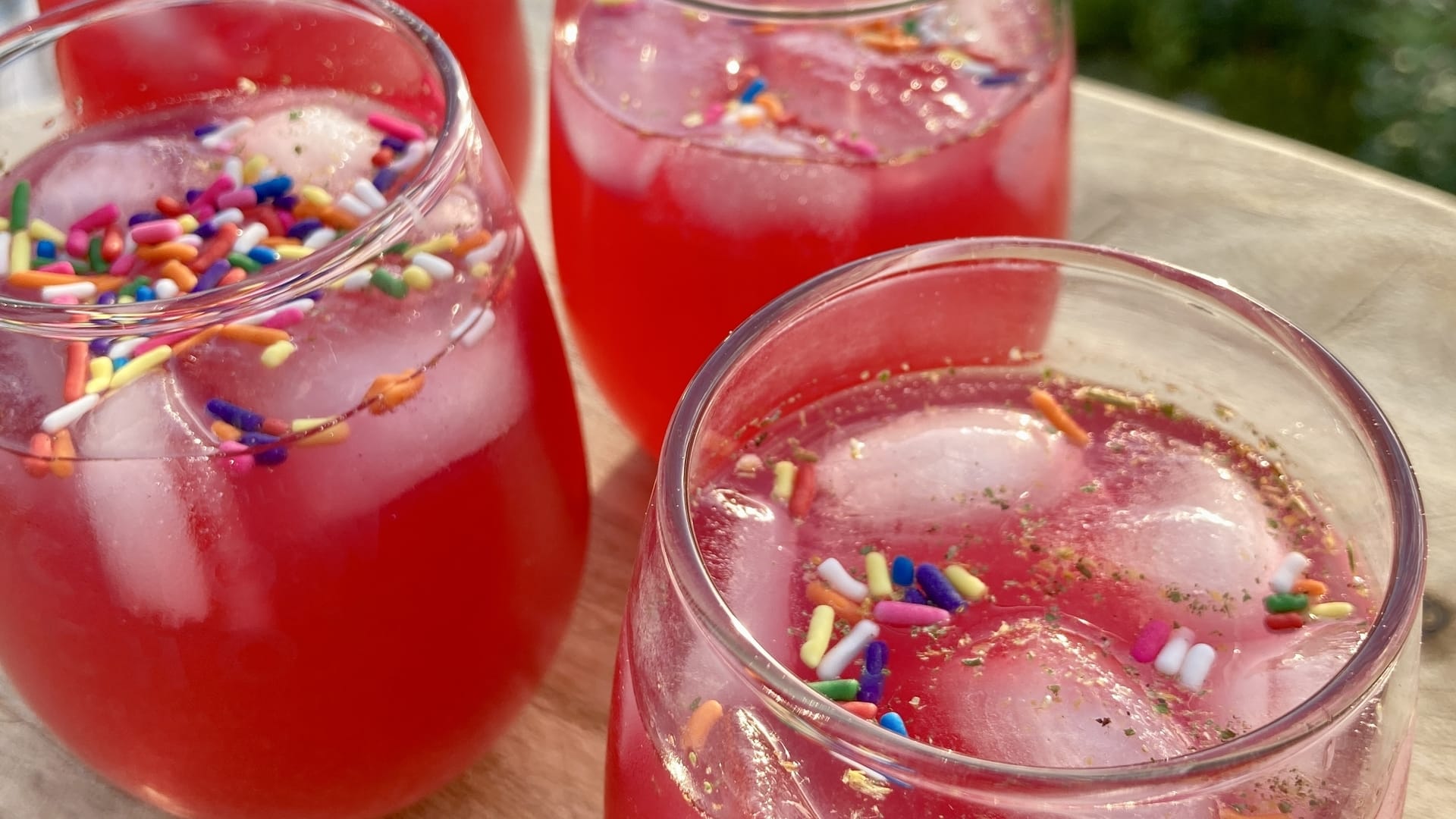 Glasses of rose colored liquid with rainbow sprinkles and ice on a wooden background. 