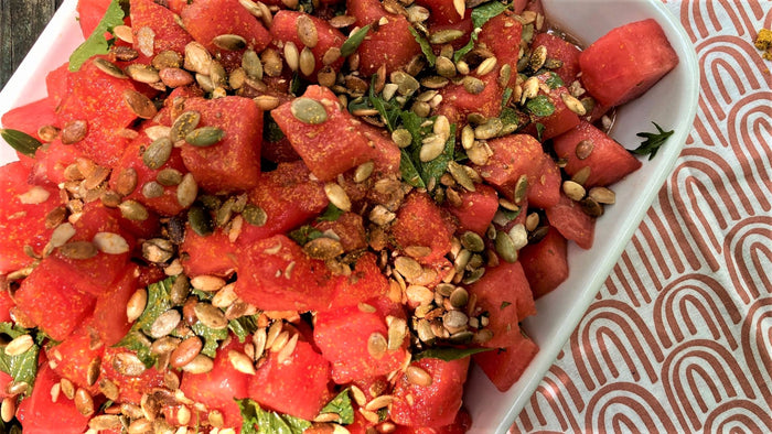 A white bowl of watermelon salad with pepitas and greens on an orange and white patterned napkin, made with Golden Milk from Well Seasoned Table
