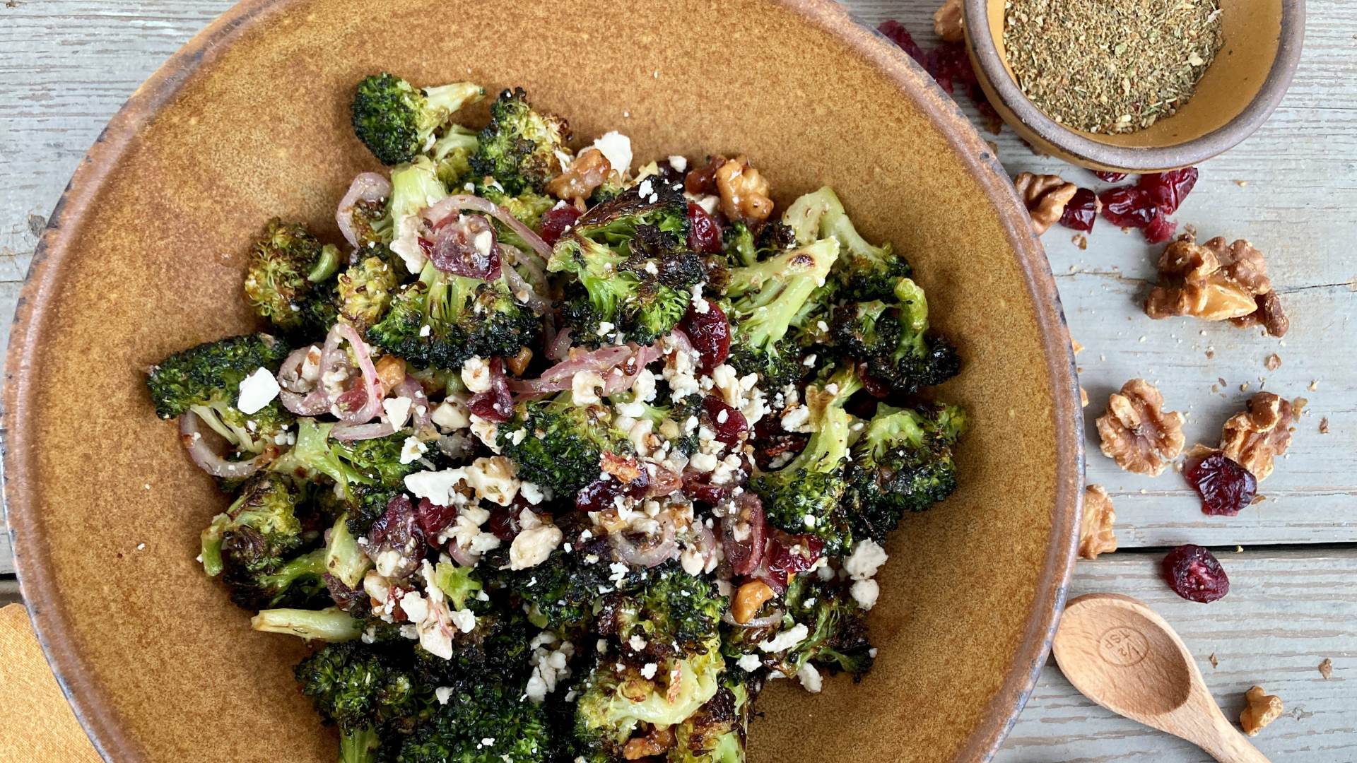 A brown ceramic bowl of Roasted Broccoli Salad on a wooden background with toasted nuts, dried cranberries, and a bowl of Wild Green Goddess Seasoning next to it.