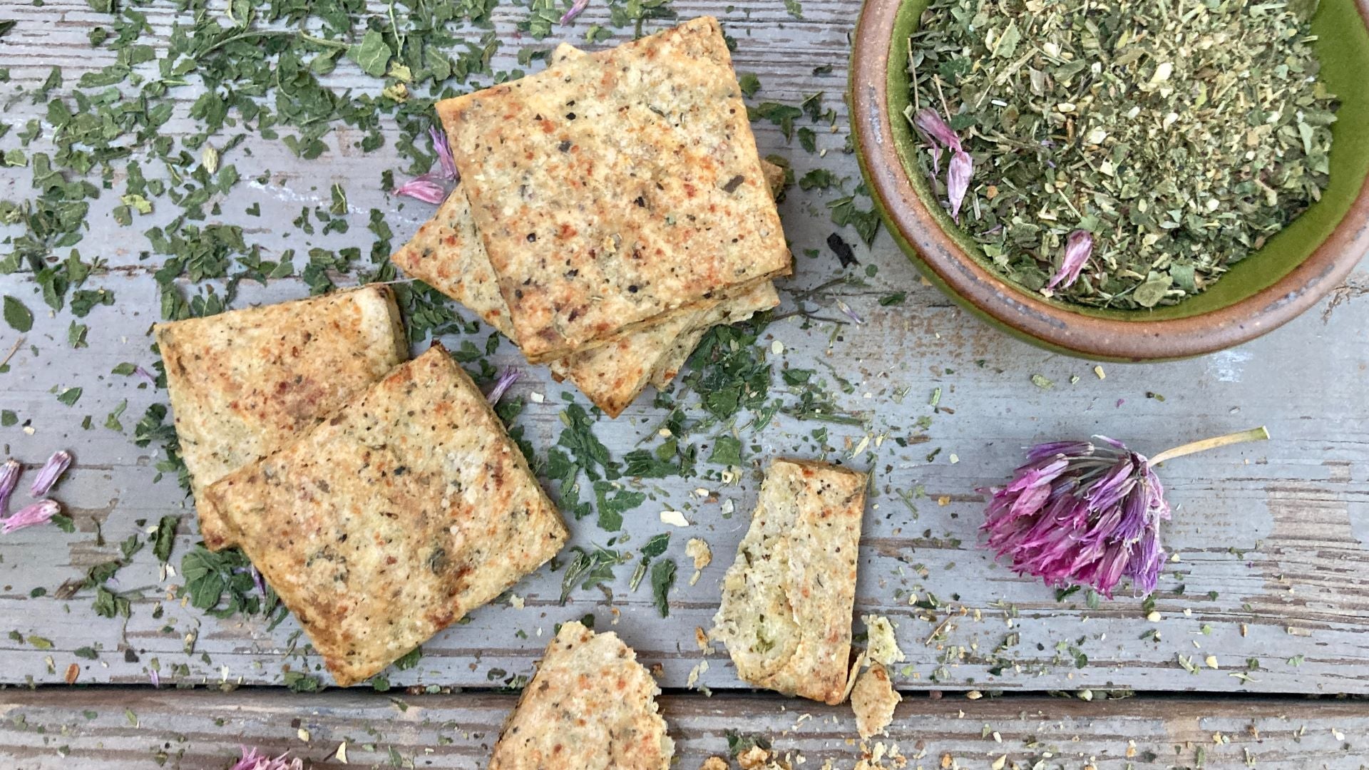Homemade crackers on a wooden background with seasoning around them and a chive blossom.