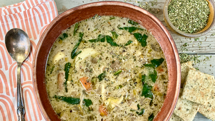 A bowl of chicken tortellini soup made with Wild Green Goddess Seasoning from Well Seasoned Table on a wooden background with a napkin, spoon, crackers, and a bowl of seasoning next to it.
