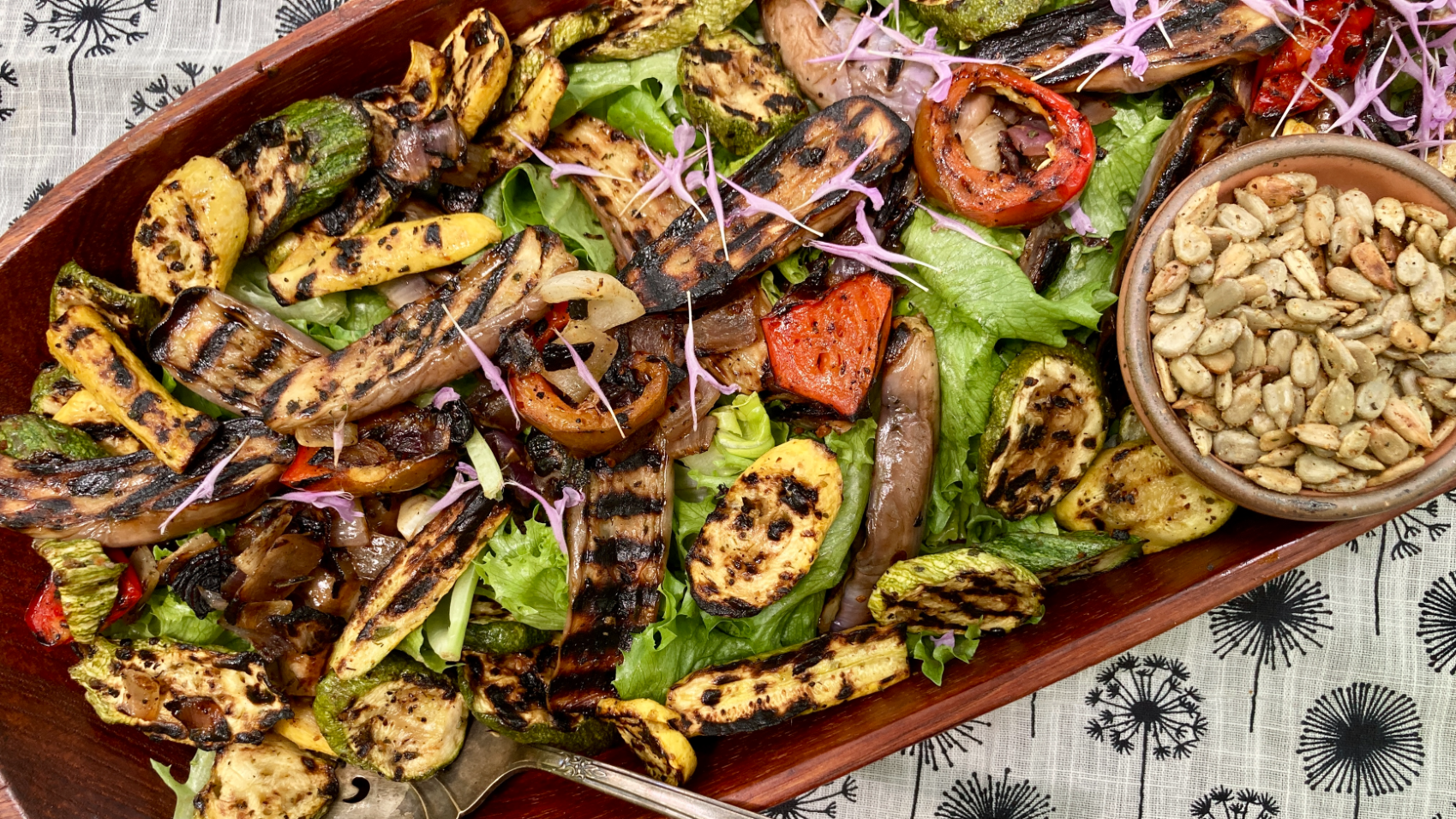 A wooden platter of girlled summer vegetables over lettuce and toasted spiced sunflower seeds on a floral textile background.