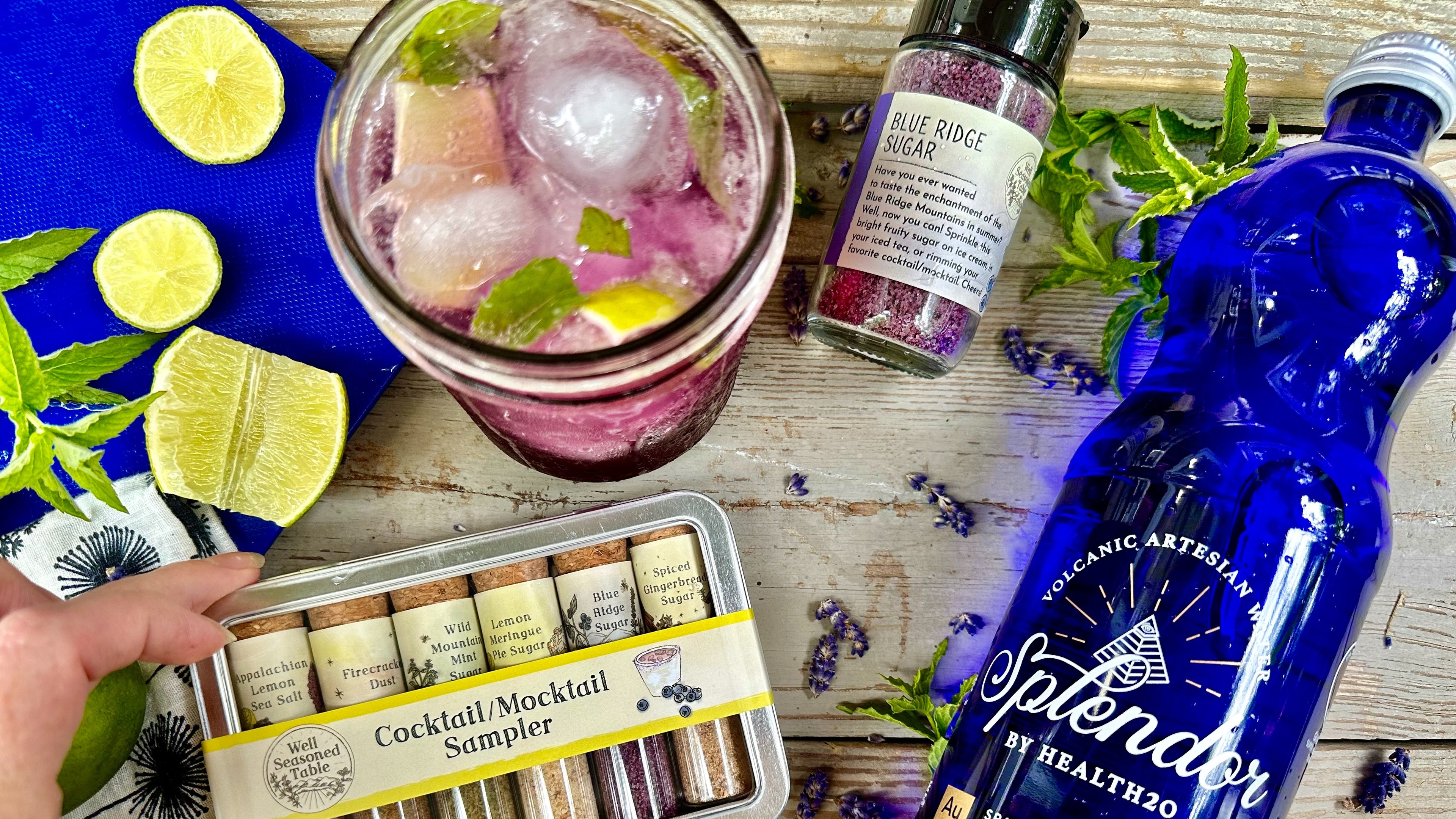 A blue bottle of Splendor sparkling water, a Cocktail/mocktail Sampler Tin, and a jar of Blue RIdge Sugar, from Well Seasoned Table with a purple cocktail, lime wedges, and mint leaves.