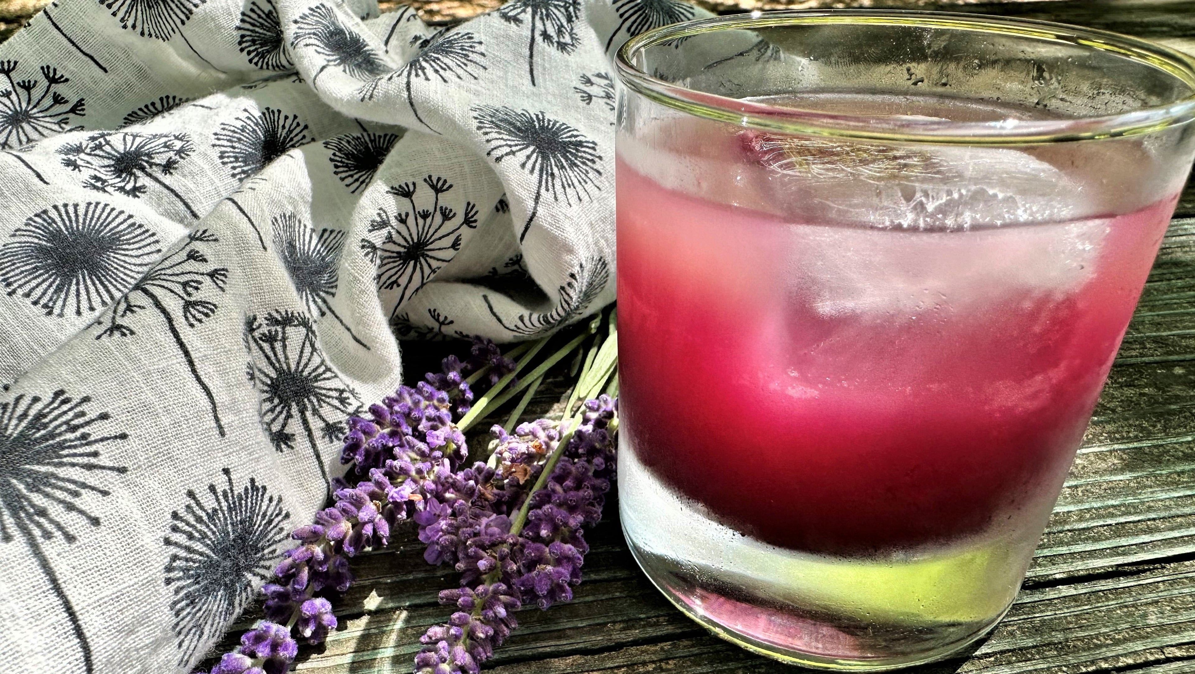 A highball glass full of bright magenta liquid and an ice cube on a wooden board with a floral napkin and fresh lavender blossoms.