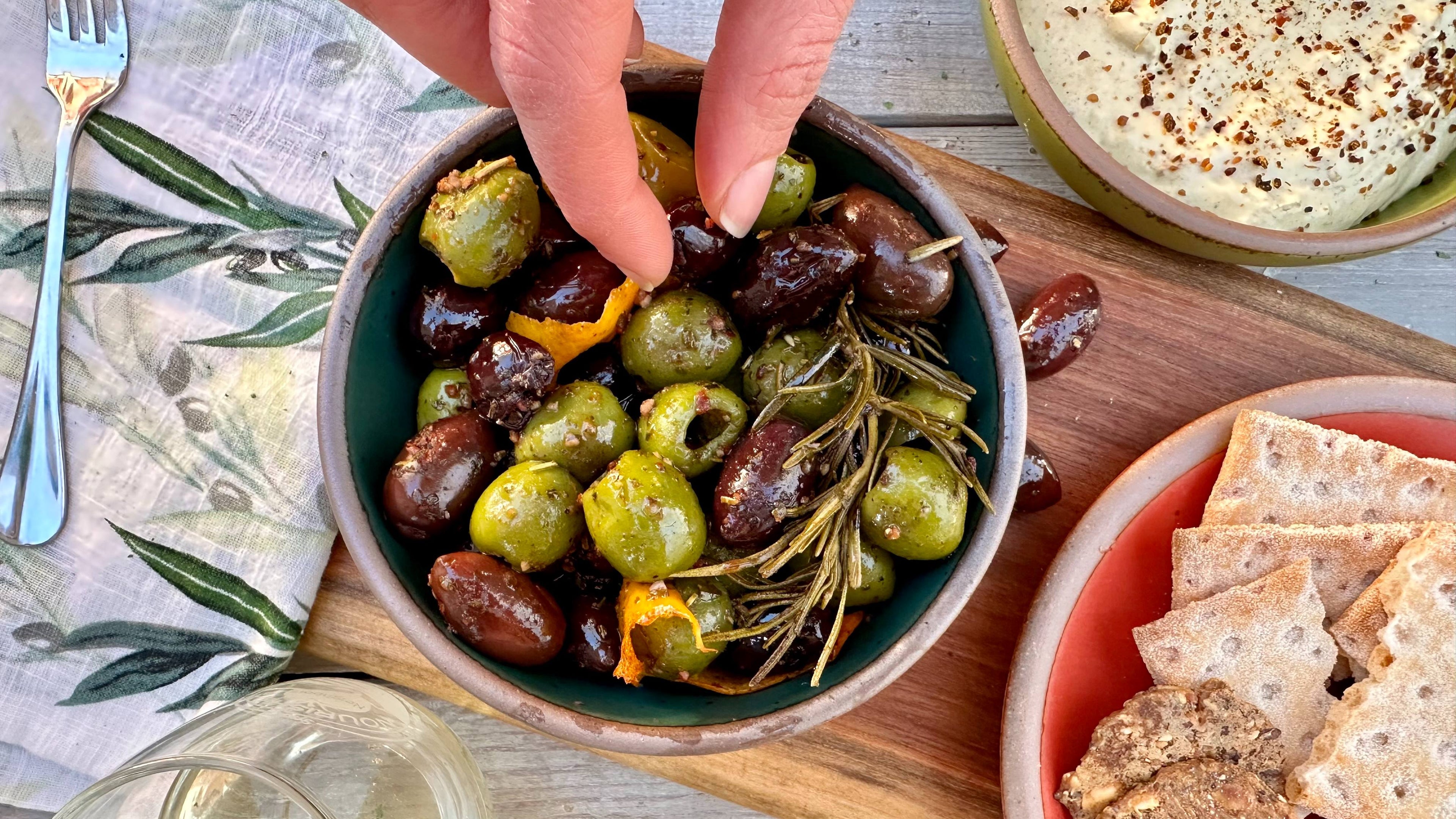 A bowl of colorful spice olives are on a charcuterie board with crackers, dip, and wine and fingers are dipping into the bowl to grab one.