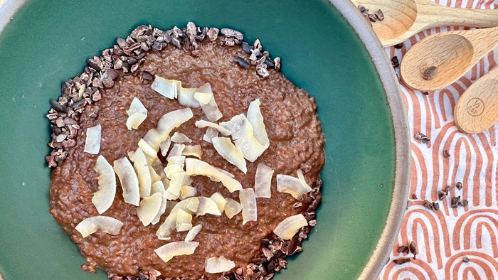 A teal ceramic bowl of chocolate chia pudding with coconut, cacao nibs, and a textile napkin behind it.