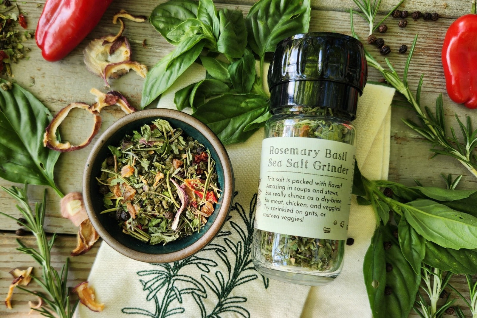 A glass grinder jar of Rosemary Basil Sea Salt from Well Seasoned Table on a wooden background with a ceramic bowl of seasoning, peppers, rosemary, and basil around it.