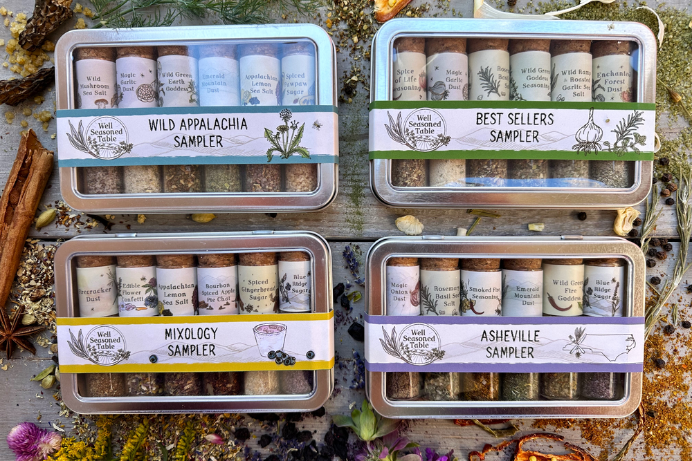 A colorful collection of metal sampler tins with 6 glass vials of infused sugars and organic farm-grown local spice blends and teas surrounded with wildcrafted flowers and dried organic herbs and spices.